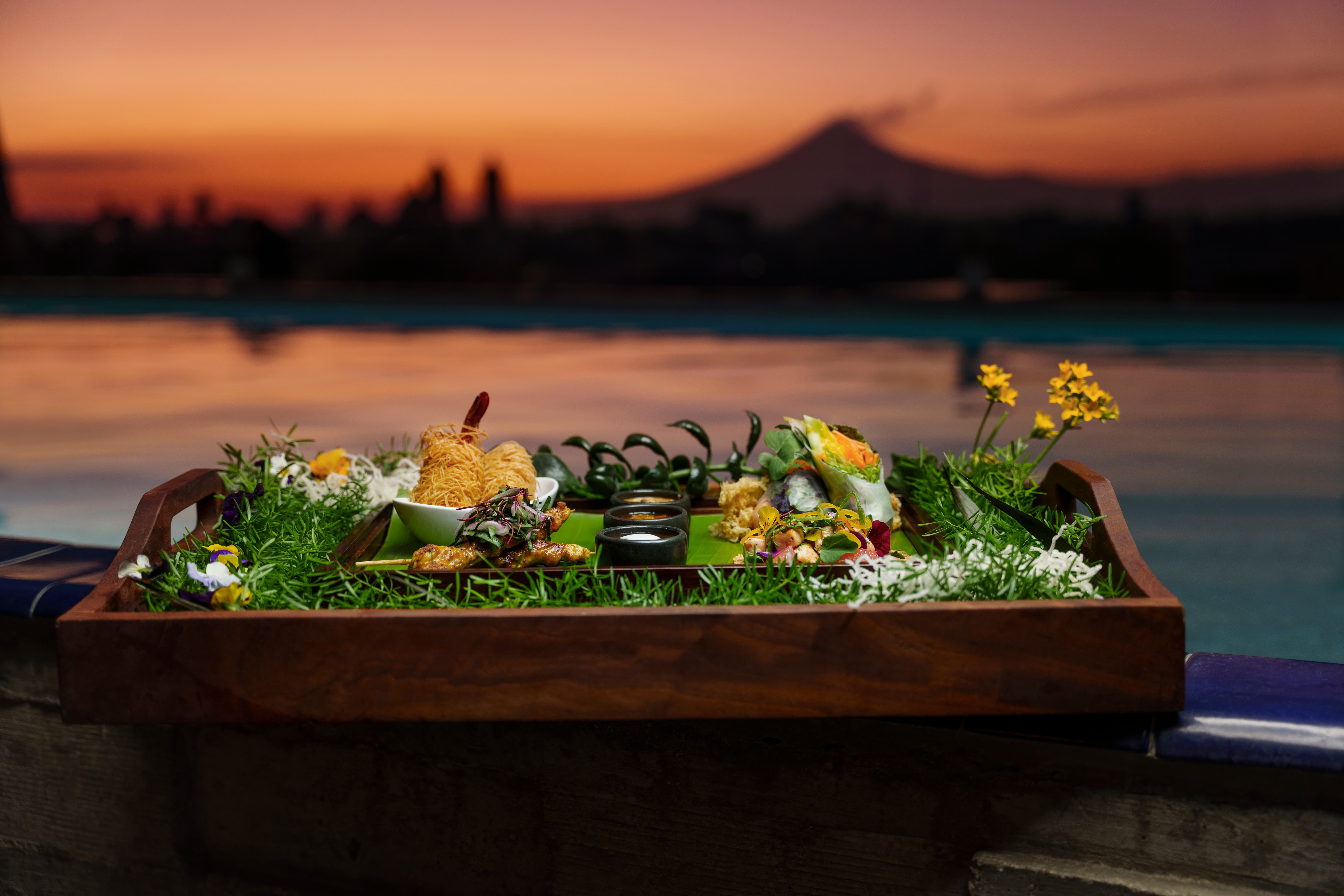 Platter of gourmet food rests by pool at dusk, overlooking Banyan Tree Puebla's tranquil scenery.
